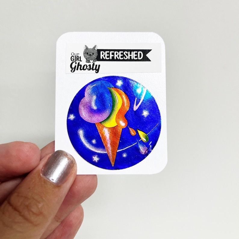 May include: A sticker with an ice cream cone illustration in vibrant colours. The ice cream is a blend of purple, yellow, and orange, set against a blue backdrop with stars and planets. The sticker also includes the text "Our Girl Ghosty" and "Refreshed".