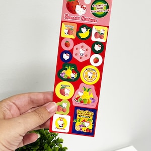 Puede incluir: Una hoja de pegatinas perfumadas Hello Kitty rojas con diseños de frutas, incluyendo fresas, plátanos y naranjas. Las pegatinas tienen el texto "Scented Stickers" y "Scratch n' Sniff Strawberry".
