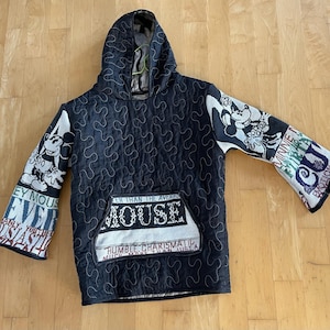 May include: Black quilted hoodie with a hood and a front pocket. The sleeves feature Mickey Mouse and Minnie Mouse designs, along with text. The pocket has the text "MOUSE" and "HUMBLE CHARISMATIC".
