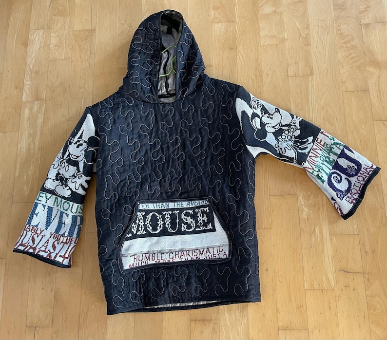 May include: Black quilted hoodie with a hood and a front pocket. The sleeves feature Mickey Mouse and Minnie Mouse designs, along with text. The pocket has the text "MOUSE" and "HUMBLE CHARISMATIC".