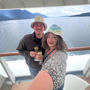 May include: A selfie of two people on a cruise ship deck, holding champagne flutes. They wear bucket hats with pastel stripes. The background shows a scenic view of mountains and water under a cloudy sky.