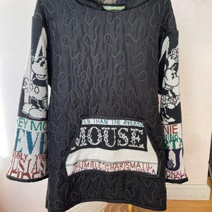 May include: Black quilted hooded jacket with white stitching and a large front pocket. The sleeves feature a Mickey Mouse design and text. The pocket has the word "MOUSE" and the phrases "HUMBLE CHARISMATIC" and "EVER THAN THE AVERAGE".