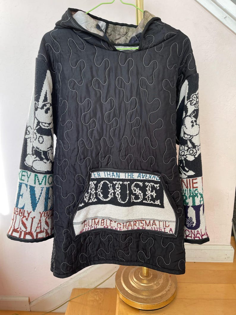 May include: Black quilted hooded jacket with white stitching and a large front pocket. The sleeves feature a Mickey Mouse design and text. The pocket has the word "MOUSE" and the phrases "HUMBLE CHARISMATIC" and "EVER THAN THE AVERAGE".