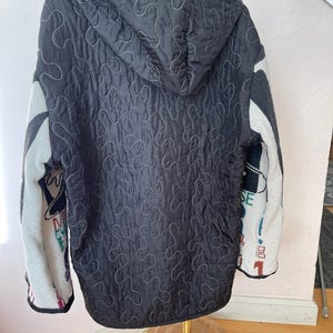 May include: Black quilted jacket with a hood and white stitching. The sleeves are a mix of gray, white, and colorful patterns. The jacket is hanging on a gold stand.