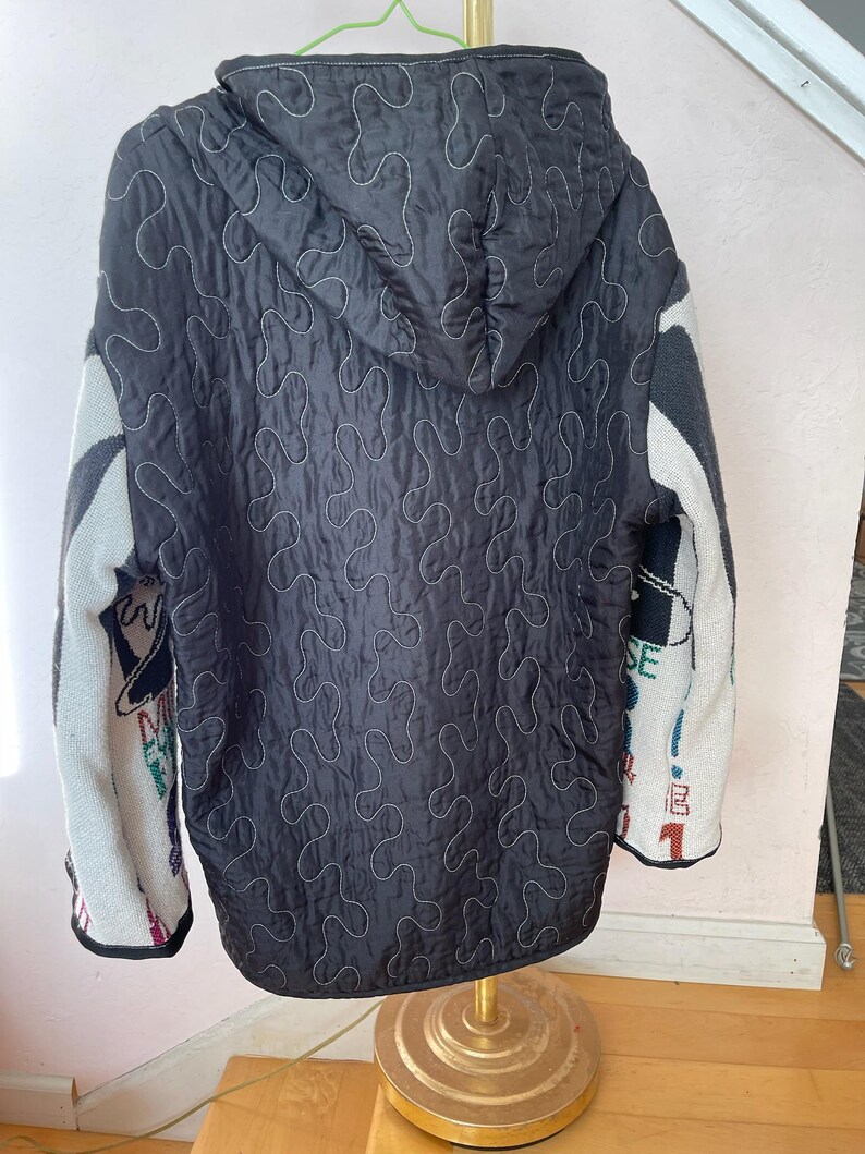 May include: Black quilted jacket with a hood and white stitching. The sleeves are a mix of gray, white, and colorful patterns. The jacket is hanging on a gold stand.