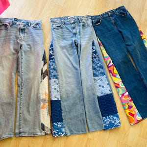 May include: Three pairs of denim jeans with unique patchwork and patterned accents. One pair is light wash with a patchwork design on the sides. Another pair is medium wash with blue patchwork. The third pair is dark wash with a colorful, psychedelic pattern.