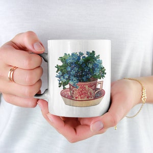 Puede incluir: Taza de cer&aacute;mica blanca con una ilustraci&oacute;n colorida de una taza de t&eacute; llena de flores azules de nomeolvides. La taza de t&eacute; est&aacute; sobre un platillo con un patr&oacute;n floral rojo y blanco.