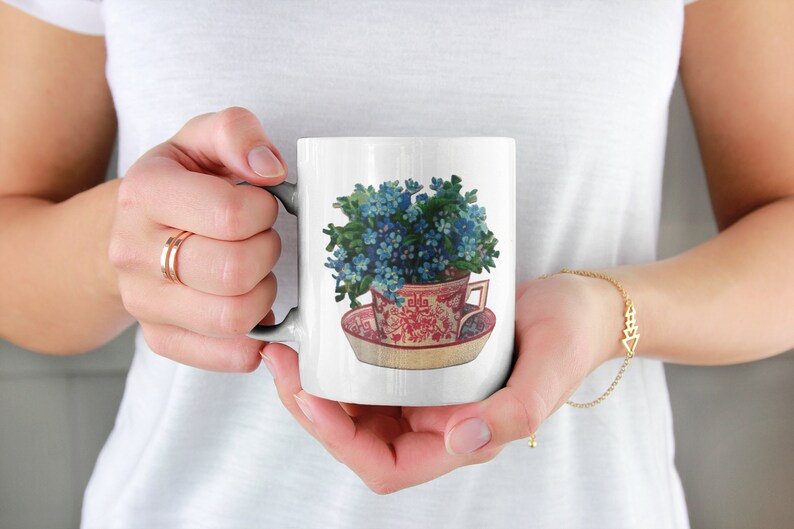 Puede incluir: Taza de cer&aacute;mica blanca con una ilustraci&oacute;n colorida de una taza de t&eacute; llena de flores azules de nomeolvides. La taza de t&eacute; est&aacute; sobre un platillo con un patr&oacute;n floral rojo y blanco.