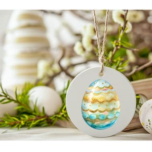 May include: White wooden Easter egg ornament with a blue and gold painted egg design inside. The ornament is hanging from a twine string.