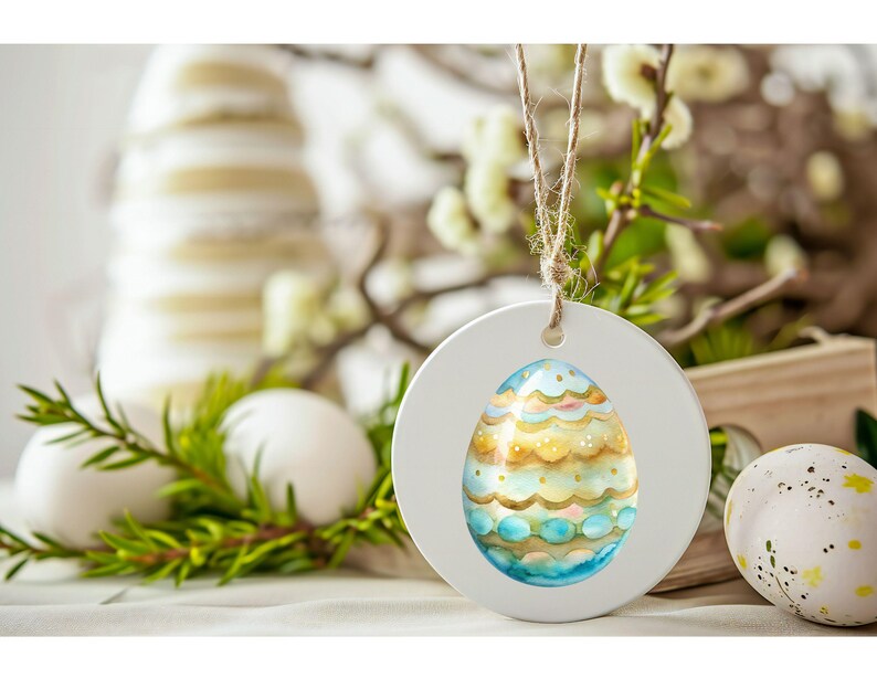 May include: White wooden Easter egg ornament with a blue and gold painted egg design inside. The ornament is hanging from a twine string.