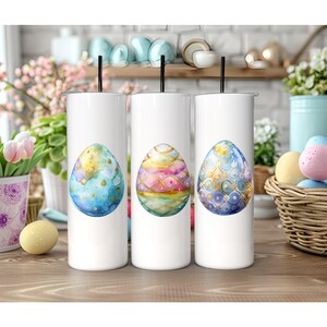 May include: Three white stainless steel tumblers with a colorful Easter egg design. Each tumbler features a different egg design with gold accents. The eggs are painted in shades of blue, pink, and purple.