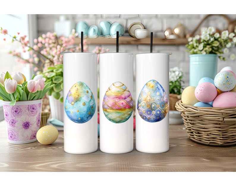May include: Three white stainless steel tumblers with a colorful Easter egg design. Each tumbler features a different egg design with gold accents. The eggs are painted in shades of blue, pink, and purple.
