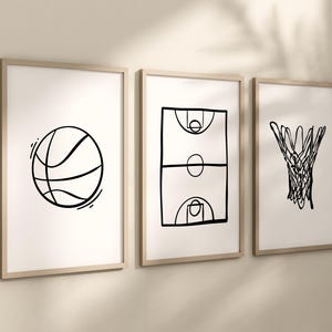 May include: Three framed art prints with basketball-themed line drawings. The prints feature a basketball, a basketball court, and a basketball net, all in black on a white background. The frames are a light wood color.