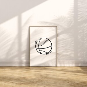 May include: A framed print featuring a minimalist black line drawing of a basketball. The artwork is displayed against a white wall with natural light and shadows, and rests on a wooden floor. The basketball design is simple and modern.