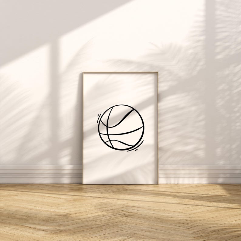 May include: A framed print featuring a minimalist black line drawing of a basketball. The artwork is displayed against a white wall with natural light and shadows, and rests on a wooden floor. The basketball design is simple and modern.