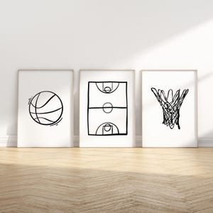 May include: Three framed art prints featuring basketball-themed line drawings. The prints depict a basketball, a basketball court, and a basketball hoop. The artwork is in black and white, with a minimalist style, and is displayed on a wooden floor.