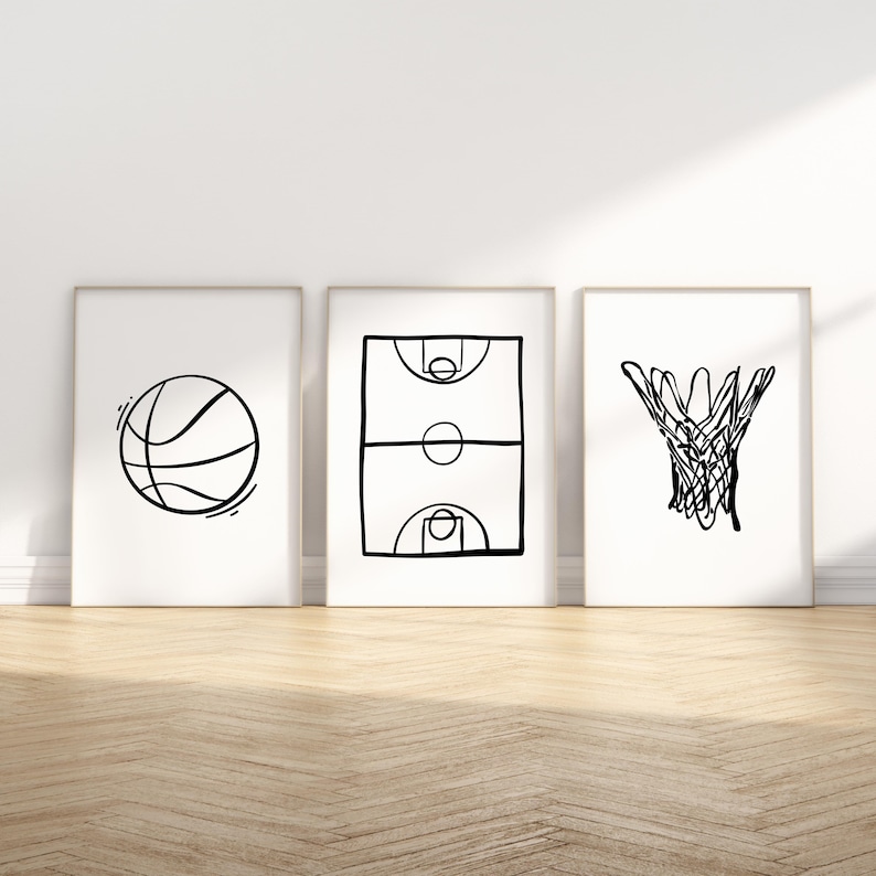 May include: Three framed art prints featuring basketball-themed line drawings. The prints depict a basketball, a basketball court, and a basketball hoop. The artwork is in black and white, with a minimalist style, and is displayed on a wooden floor.