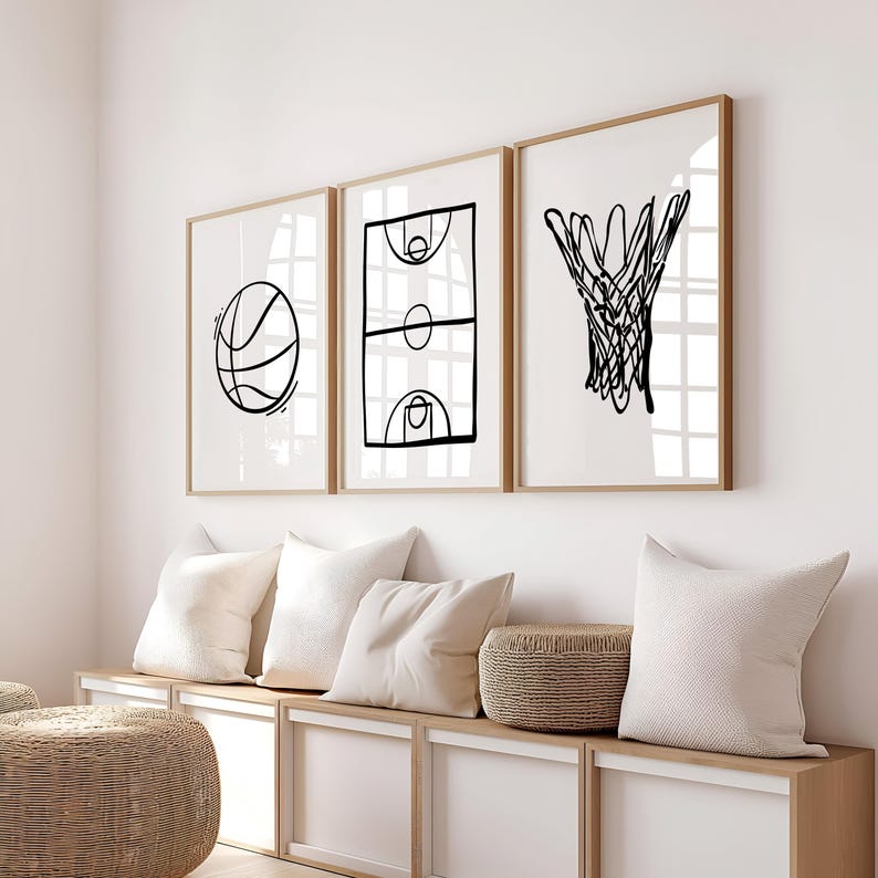 May include: Three framed art prints with basketball-themed line drawings. The prints feature a basketball, a basketball court, and a basketball net. The frames are light brown, and the background is white. The prints are displayed above a white storage bench with pillows.