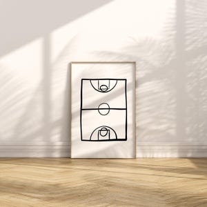 May include: A framed minimalist art print featuring a black line drawing of a basketball court. The artwork depicts the court's key elements: the hoops, the center circle, and the free-throw lanes. The print is displayed against a white wall with a wooden floor.