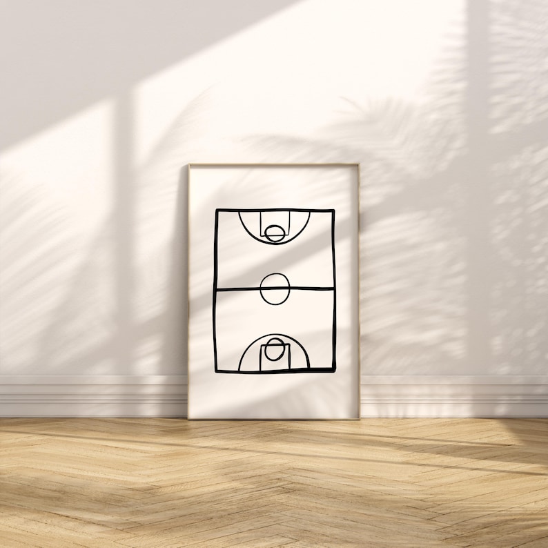 May include: A framed minimalist art print featuring a black line drawing of a basketball court. The artwork depicts the court's key elements: the hoops, the center circle, and the free-throw lanes. The print is displayed against a white wall with a wooden floor.