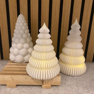 Könnte beinhalten: Drei weiße dekorative Weihnachtsbaumfiguren. Einer hat eine strukturierte, blasenartige Oberfläche, während die anderen beiden ein geschichtetes, geripptes Design haben. Sie werden auf einer kleinen Holzpalette vor einem hölzernen Lattenhintergrund ausgestellt.