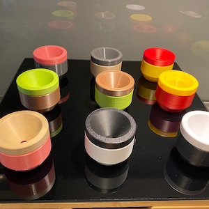 Könnte beinhalten: Eine Ansammlung von neun kleinen, zweifarbigen Kunststoffschalen, die in einem Raster angeordnet sind. Jede Schale weist eine eigene Farbkombination auf, darunter Rosa, Limettengrün, Beige, Grau, Rot, Gelb, Orange, Schwarz und Weiß mit trichterförmigem Innenraum.