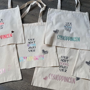 Könnte beinhalten: Sechs weiße Stofftaschen mit dem Text "VIER ACHT SECHS ZWEI VIER SCHÖPPINGEN" auf jeder Tasche. Der Text ist auf jeder Tasche in verschiedenen Farben und Schriftarten.