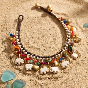 May include: A dark brown braided cord anklet featuring a variety of charms including white carved elephant figures, gold-toned hearts, small metallic bells, and colourful beads in red, blue, orange, and green, arranged along the length of the cord.