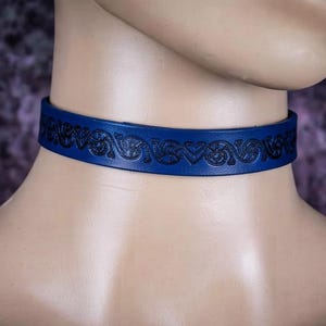 Op de afbeelding: Een blauwe leren choker met een doorlopend, in reliëf aangebracht zwart patroon van herhalende krullen en hartvormige figuren. De band is glad en plat, omsluit de nek met een constante breedte en een donker, gedetailleerd decoratief ontwerp.