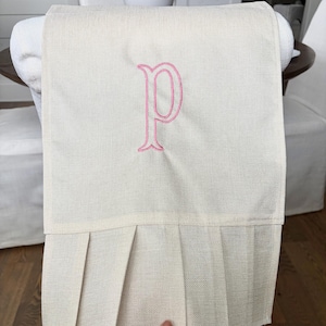 Puede incluir: Funda de silla color crema con una falda plisada y una letra "P" bordada en rosa. La funda está hecha de una tela texturizada y está colocada sobre el respaldo de una silla.