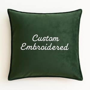 Op de afbeelding: Een vierkant, donkergroen fluwelen kussen met een contrasterende donkergroene rand. De woorden "Custom Embroidered" zijn in witte letters over het midden gestikt en bieden een persoonlijk tintje. Het kussen is een decoratief woonaccessoire.