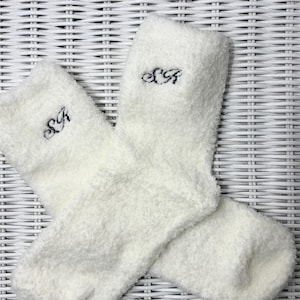 以下が含まれることがあります： 黒い糸で「S A」のイニシャルが刺繍された、白くふわふわの靴下のペア。 靴下は柔らかく、質感のある素材でできており、折り返されています。 背景は白い籐の模様です。