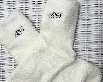 Puede incluir: Un par de calcetines blancos y esponjosos con las iniciales "S A" bordadas en hilo negro. Los calcetines están hechos de un material suave y texturizado y están doblados. El fondo es un patrón de mimbre blanco.