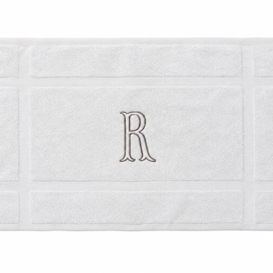 May include: A rectangular white bath mat featuring a textured border design and a centered, embroidered letter R in a light gray thread. The mat has a soft, plush surface with a distinct frame pattern surrounding the central monogrammed area.