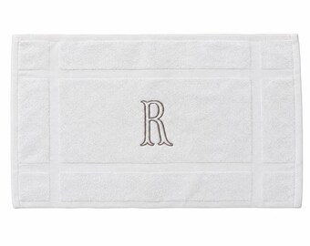 May include: A rectangular white bath mat featuring a textured border design and a centered, embroidered letter R in a light gray thread. The mat has a soft, plush surface with a distinct frame pattern surrounding the central monogrammed area.