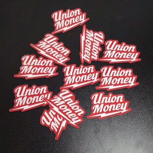 May include: A collection of red and white stickers with the words "Union Money" in a stylized font. The stickers have a lightning bolt design element and are scattered on a dark surface.