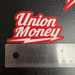 May include: A red and white sticker with the words "Union Money" in a stylized font. A lightning bolt design is at the bottom. A ruler is placed next to the sticker for scale.