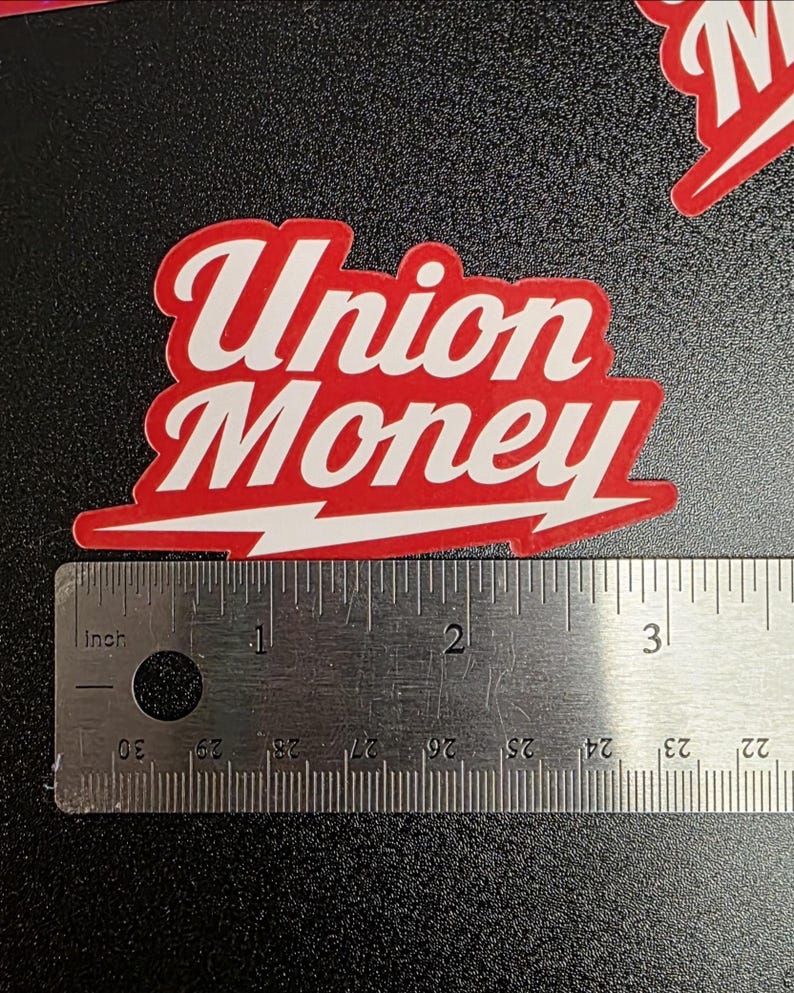 May include: A red and white sticker with the words "Union Money" in a stylized font. A lightning bolt design is at the bottom. A ruler is placed next to the sticker for scale.