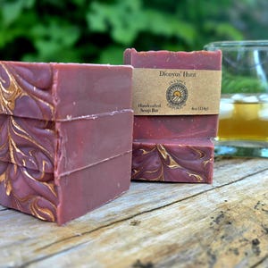 May include: Stacked bars of handcrafted soap with a burgundy hue and gold decorative swirls. One bar is labeled "Dionysus' Hunt" with a logo and weight. A glass of amber liquid with ice sits nearby on a wooden surface.