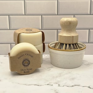 Op de afbeelding: Een stapel van drie ronde, gebroken witte afwaszeepblokken verpakt in bruine papieren labels met de tekst Sweet Orange Dish Soap Refill, naast een houten afwasborstel met stijve haren in een kleine, witte keramische kom met een bruine rand.