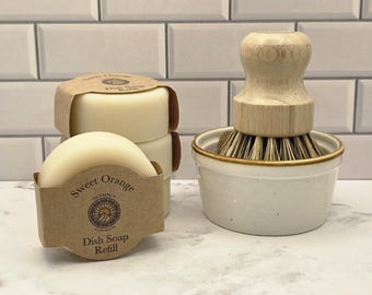 Può includere: Una pila di tre panetti di sapone per piatti rotondi color bianco sporco avvolti in etichette di carta marrone con la scritta Sweet Orange Dish Soap Refill, accanto a una spazzola per piatti in legno con setole rigide in una ciotola di ceramica.