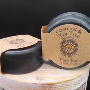 May include: Two round black charcoal and tea tree face bars with brown paper labels. The label on the front bar reads "Charcoal & Tea Tree Olympus Emporium Face Bar 2.85oz (79g)".
