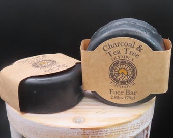 Puede incluir: Dos barras faciales redondas de color negro con carbón y árbol de té, envueltas en bandas de papel color canela. Las etiquetas muestran el texto Charcoal & Tea Tree, Olympus Emporium y Face Bar 2.85oz (79g) con un logotipo circular.