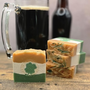 May include: A stack of handmade soaps with green and orange swirls, and a single bar with a green and white design. A glass of dark beer and a bottle are in the background. The soaps are on a wooden surface.