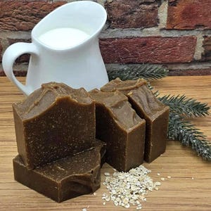 Può includere: Tre barre rettangolari di sapone marrone scuro testurizzato con particelle di avena visibili, disposte vicino a una brocca in ceramica bianca riempita di latte e un piccolo mucchio di avena cruda, per un aspetto rustico e naturale.