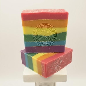 Peut inclure: Deux pains de savon rectangulaires empilés verticalement, présentant des rayures horizontales rouges, orange, jaunes, vertes, bleues et violettes. Le pain supérieur porte un tampon circulaire en relief représentant un soleil levant sur des montagnes.