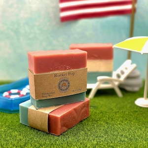 May include: Two rectangular soap bars stacked, featuring red, white, and blue layers. A brown paper label wraps around the center, printed with 'Rocket Pop', 'Olympus Emporium', and '4oz (114g)'. The soap has a smooth texture and a stamped logo.