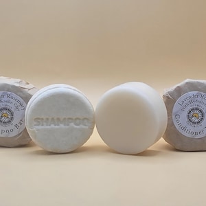 May include: Four natural hair care bars. Two round shampoo bars, one with "SHAMPOO" embossed, and two wrapped bars labeled "Lavender Rosemary" with "Shampoo Bar" and "Conditioner Bar". The bars are light beige.
