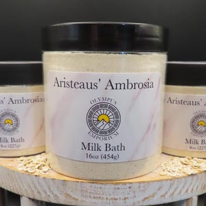 Op de afbeelding: Drie potten met witte badschuim, gelabeld "Aristeaus' Ambrosia" met de tekst "Olympus Emporium" en "Melkbad" op de voorkant. De potten staan op een wit houten oppervlak met havervlokken eromheen.