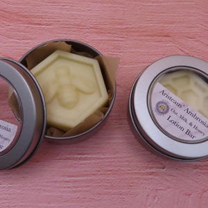 Op de afbeelding: Twee zilveren blikken met doorzichtige deksels die een witte lotionbar in de vorm van een honingraat met een bijenontwerp bevatten. Het etiket op de blik staat "Aristeaus' Ambrosia Oat, Milk & Honey Lotion Bar".